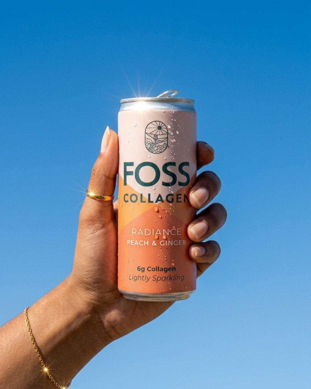 A hand holding a can of Foss Collagen Radiance Peach & Ginger sparkling drink against a clear blue sky.