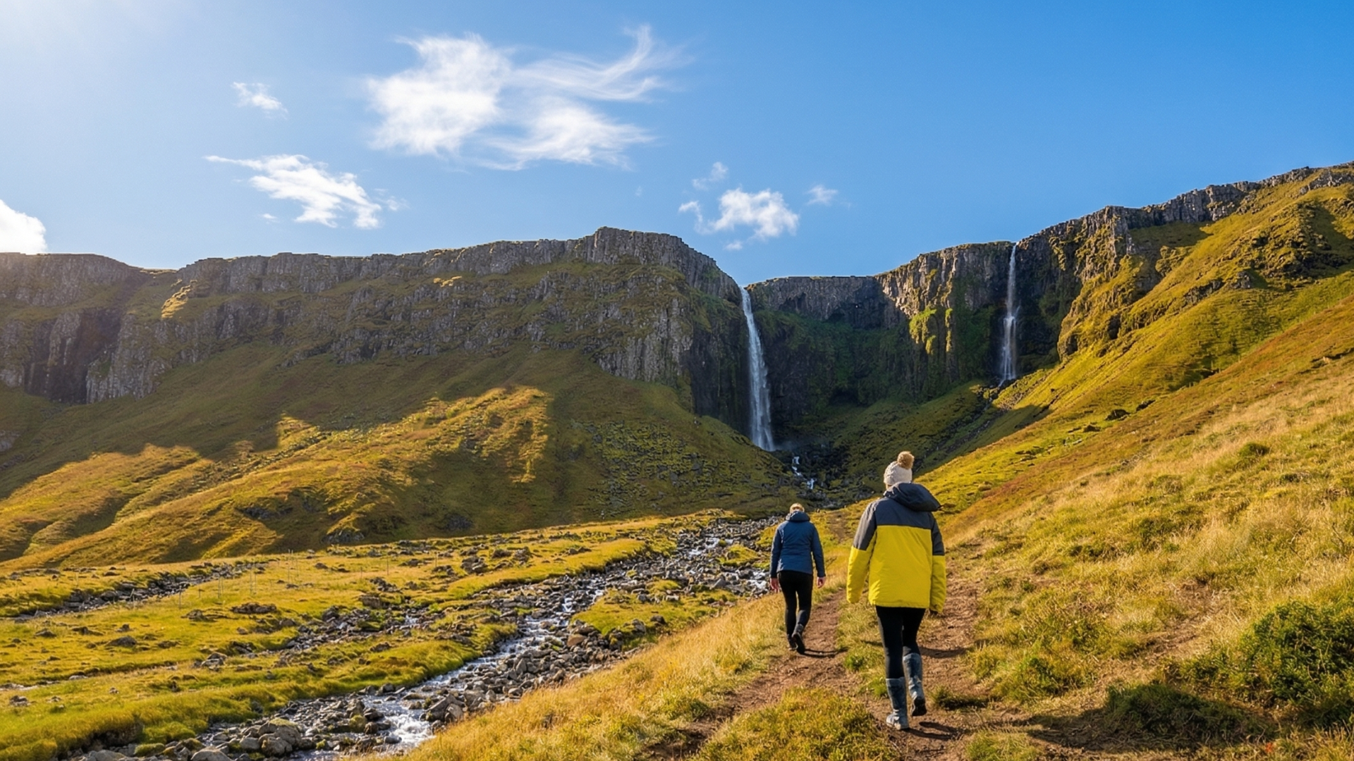 Two people hiking on a grassy path toward a tall waterfall in a dramatic mountain landscape under a blue sky with scattered clouds.